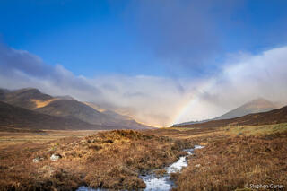 Rainbow arching over a rocky creek in the Scottish Highlands, with dark clouds and green hills in the background.